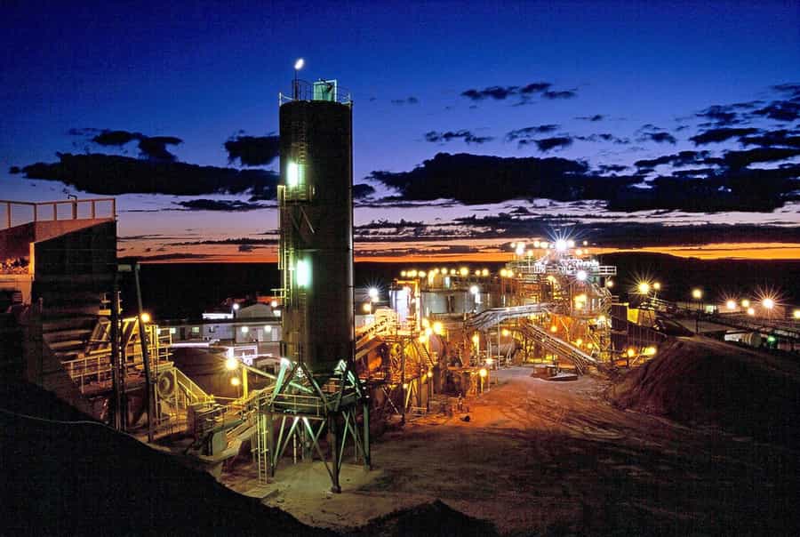 Gold Mining Facility Illuminated at Night Night view of a gold mining facility, brightly lit against a dark sky with sunset hues in the background, showcasing industrial structures and machinery involved in gold processing.